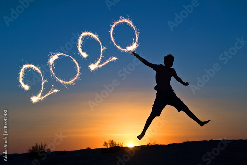 2020 written with sparkles, silhouette of a boy jumping in the sun, holiday card © Delphotostock