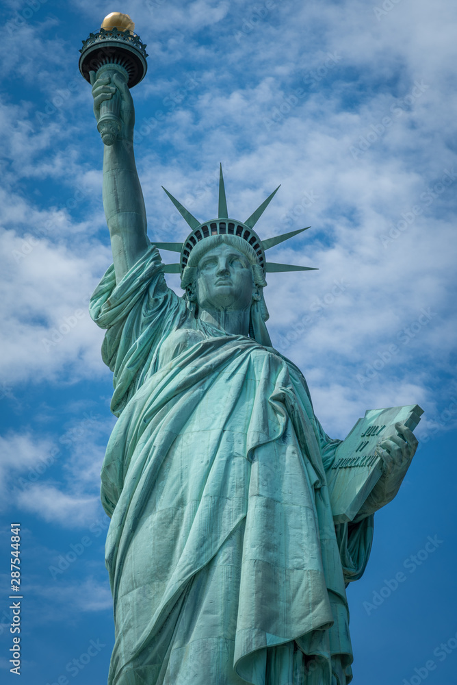 Fototapeta premium Ujęcie Statuy Wolności w Nowym Jorku, USA. Zdjęcie zostało zrobione podczas pięknego, słonecznego dnia, na tle błękitnego nieba i białych chmur