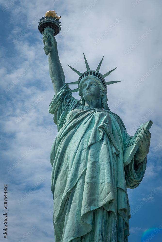 Fototapeta premium Ujęcie Statuy Wolności w Nowym Jorku, USA. Zdjęcie zostało zrobione podczas pięknego, słonecznego dnia, na tle błękitnego nieba i białych chmur