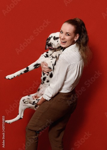 Cute happy woman in white shirt and pants, is holding dalmatian dog in her arms on red background. Love to the animals, pets friendship concept. Copy space