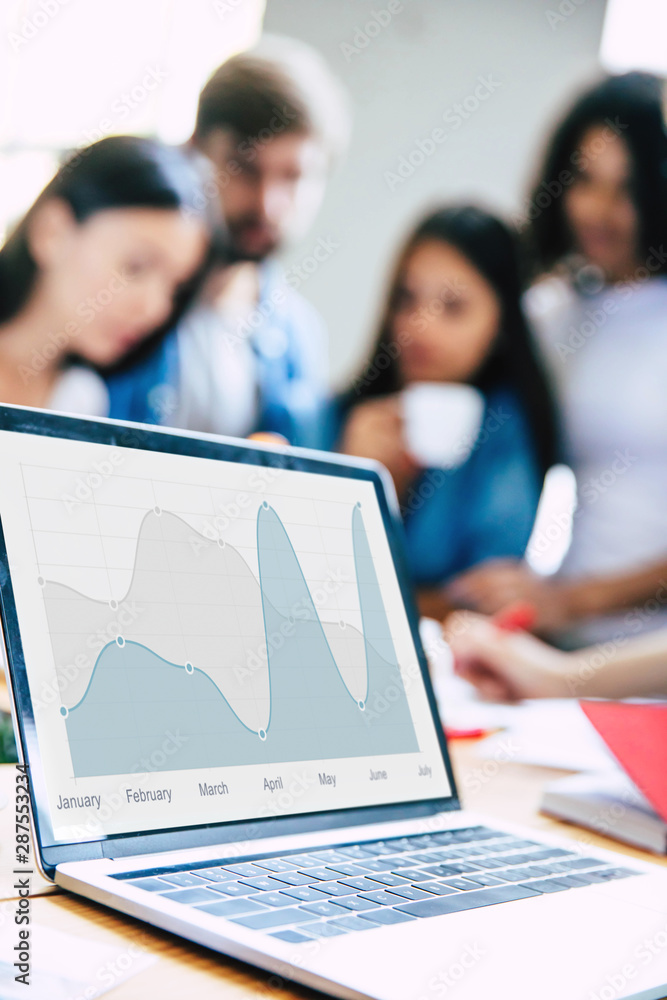 Modern laptop with diagram or graph on screen stands on table on business start up team background Modern laptop with diagram or graph on screen stands on table on business start up team background