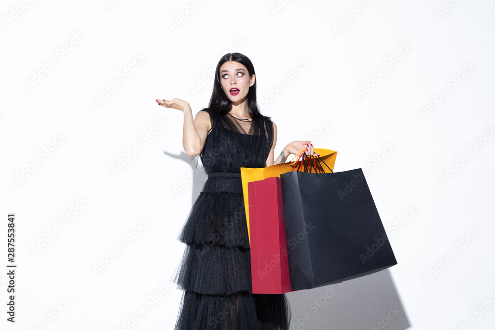 Young brunette woman in black dress shopping on white background. Attractive caucasian female model. Finance, black friday, cyber monday, sales, autumn concept. Copyspace. Pointing, showing.