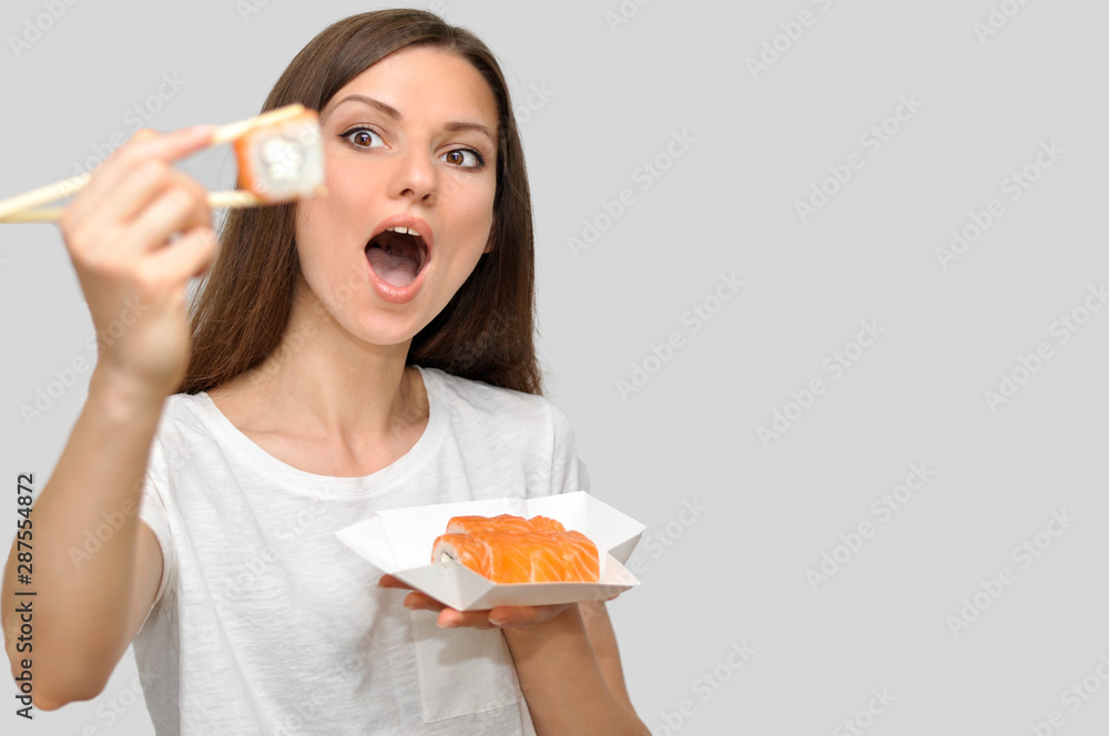 A beautiful young woman in a white T-shirt is smiling and holding a box of rolls of food delivery.A woman holds a roll near her mouth and is going to eat it. A beautiful young woman in a white T-shirt is smiling and holding a box of rolls of food delivery.A woman holds a roll near her mouth and is going to eat it.