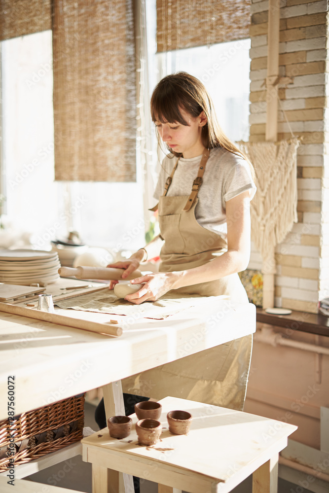 Beautiful young woman making ceramic ware, roll out with a rolling pin in sun light. Concept for woman in freelance, business, hobby Beautiful young woman making ceramic ware, roll out with a rolling pin in sun light. Concept for woman in freelance, business, hobby