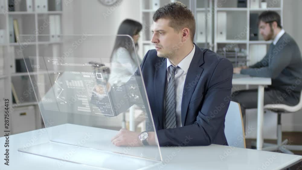 Businessman with bionic prosthetic hand sitting at desk in the office, tapping and swiping on augmented reality computer screen while working with financial data Businessman with bionic prosthetic hand sitting at desk in the office, tapping and swiping on augmented reality computer screen while working with financial data