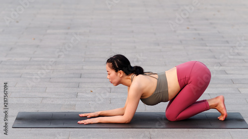 Side view of healthy women in sportswear, red pants and top practicing yoga outdoor, kneeling on yoga mat and relaxing her body.
