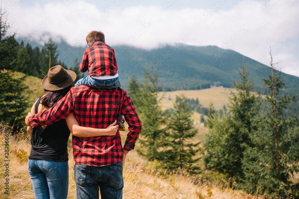 Travelers in checkered red shirts walk by the mountains. Dad and son enjoy the view of the mountains and forest. The child sits on the shoulders of his father