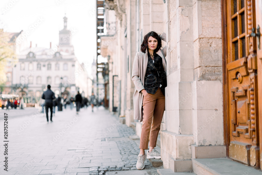 Outdoor portrait of high fashion female model in stylish clothes, coat and pants, posing in the old city street