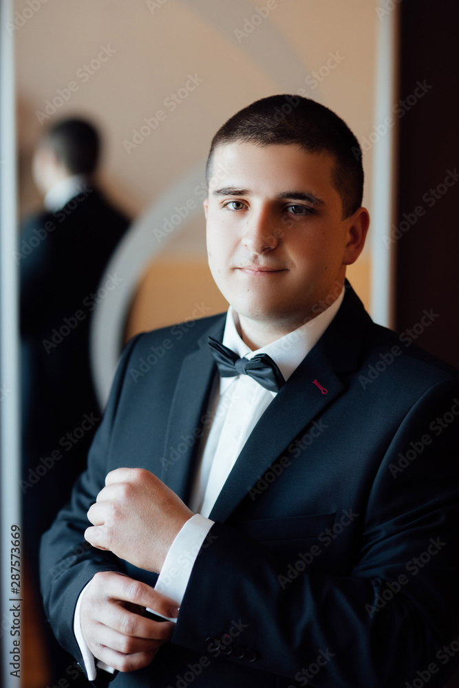 Portrait handsome groom in suit near mirror. Handsome groom on his wedding day. The groom is dressed at home in a suit. Stylish groom portrait getting ready in the morning for wedding ceremony. Portrait handsome groom in suit near mirror. Handsome groom on his wedding day. The groom is dressed at home in a suit. Stylish groom portrait getting ready in the morning for wedding ceremony.