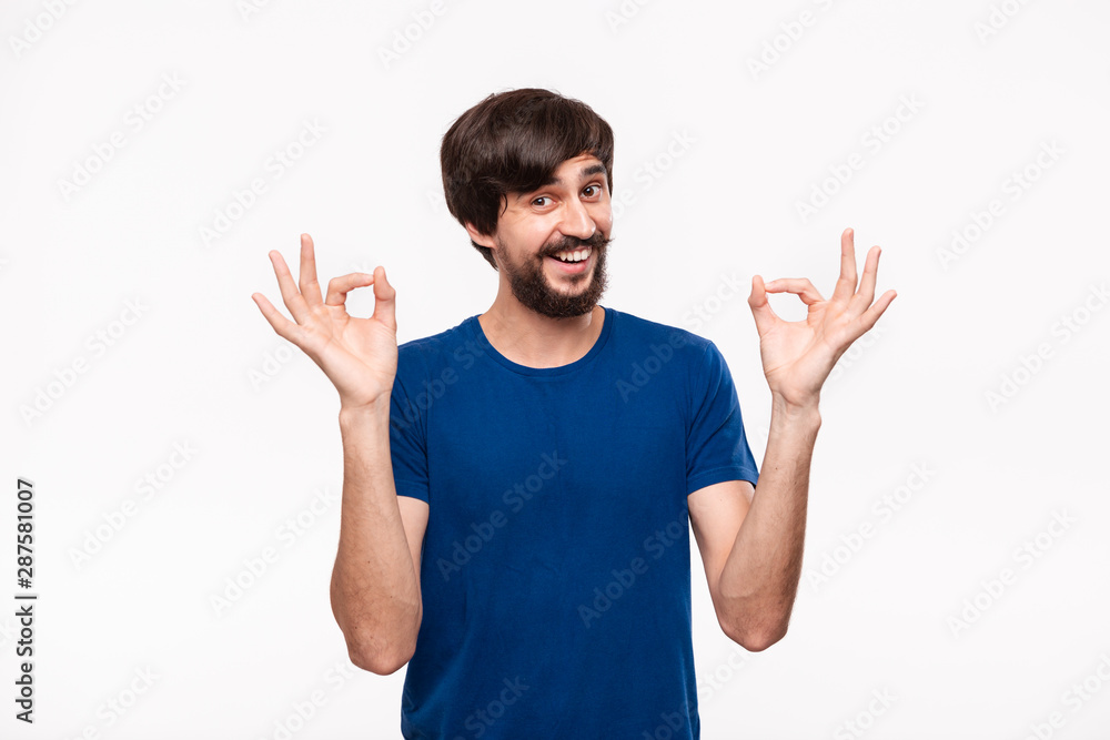 Handsome brunet bearded man with mustaches in a blue shirt showing gesture of OK sign with two hands standing isolated over white background. Handsome brunet bearded man with mustaches in a blue shirt showing gesture of OK sign with two hands standing isolated over white background.
