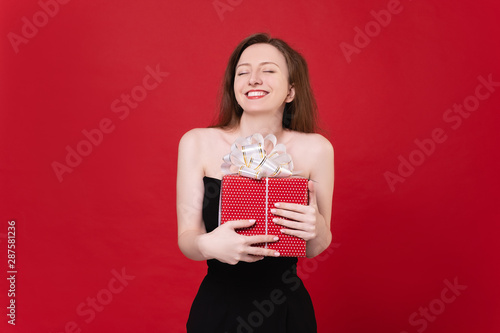 Beautiful luxurious young woman in little black dress joyfully clutching gift in red packaging posing on red background. Concept of personal offers of discounts and promotions. Advertising space © Rithor