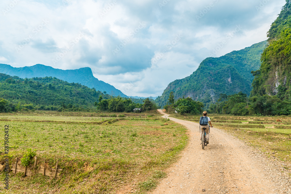 Woman riding mountain bike on dirt road in scenic landscape around Vang Vieng backpacker travel destination in Laos Asia rock pinnacles green valley