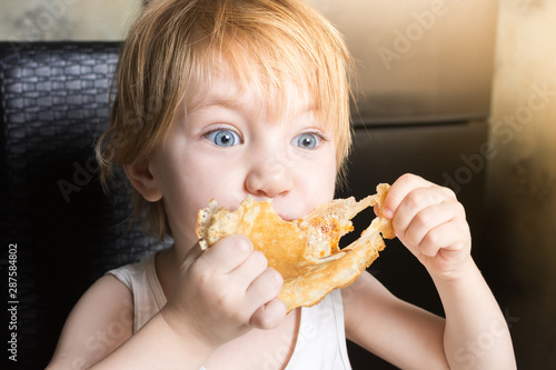 Beautiful baby with blue eyes eats delicious pancakes while sitting at kitchen table on chair.
