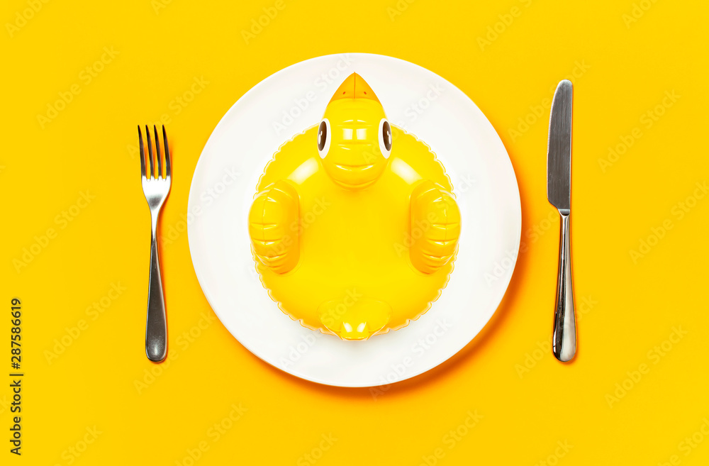 Inflatable mini yellow chicken or duckling on white plate, fork knife on yellow background. Creative food concept, tobacco chicken. Trend Inflatable Children toy for swimming. Layout for design Inflatable mini yellow chicken or duckling on white plate, fork knife on yellow background. Creative food concept, tobacco chicken. Trend Inflatable Children toy for swimming. Layout for design