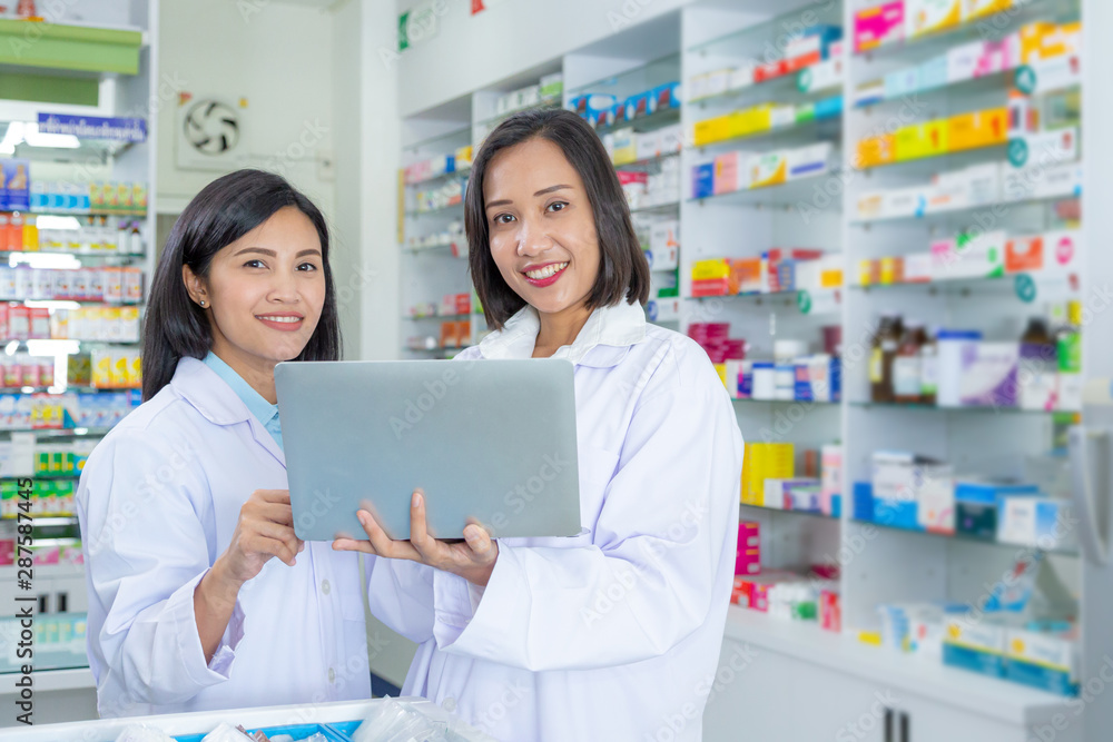 Two Asian pharmacists are using a laptop for work in a pharmacy. Health care and medical concept.