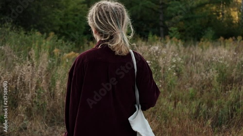 Girl walks through the meadow in the high grass at the end of the summer at cold summer day. Trendy hipster girl with dreadlocks enjoying nature back view. photo