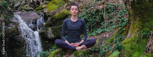 Young slim woman practicing yoga outdoors in moss forest on background of waterfall. Unity with nature concept. Girl meditates sitting