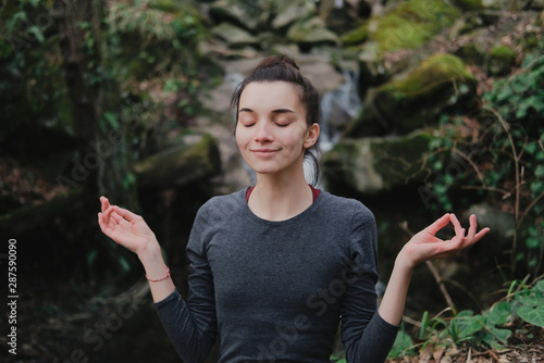 Young woman practicing breathing yoga pranayama outdoors in moss forest on background of waterfall. Unity with nature concept.