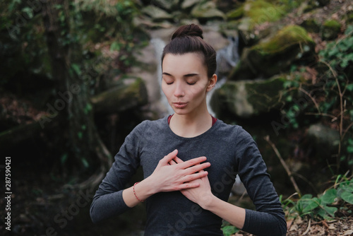 Young woman practicing breathing yoga pranayama outdoors in moss forest on background of waterfall. Unity with nature concept.