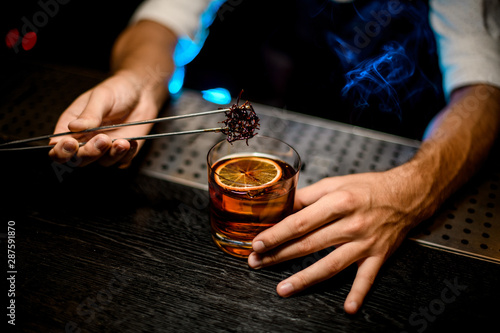 Bartender adding chilled melting caramel with twezzers to the cocktail with dried orange under blue light and smoke