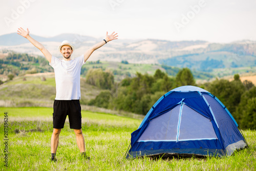 Young man with hands up camping tent facing amazing views of mountain with blue sky.