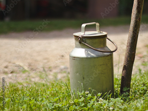 An old milk churn standing outside on the grass next to a stick