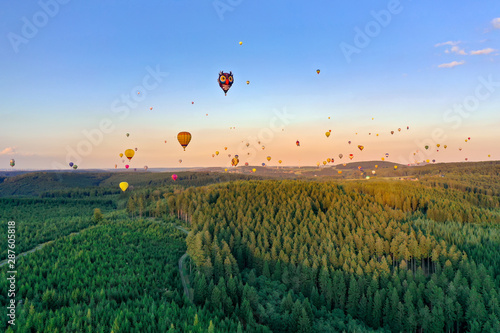 Colorful hot air balloons in different shapes fly over the forest. It is a coniferous forest in the Sauerland. The sun is almost setting, the sky is blue. Aerial shot.