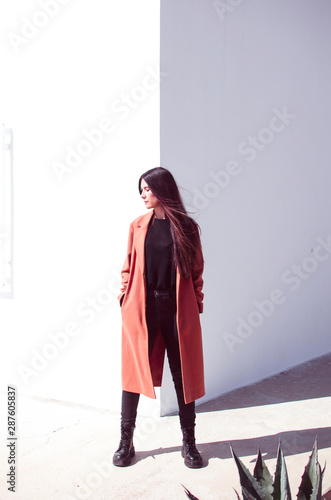 Young long-haired woman is wearing a trench coat standing in front of a corner of a wall where one side is lit and the other in the shade. She has her face turned towards sunlight to enjoy the sun