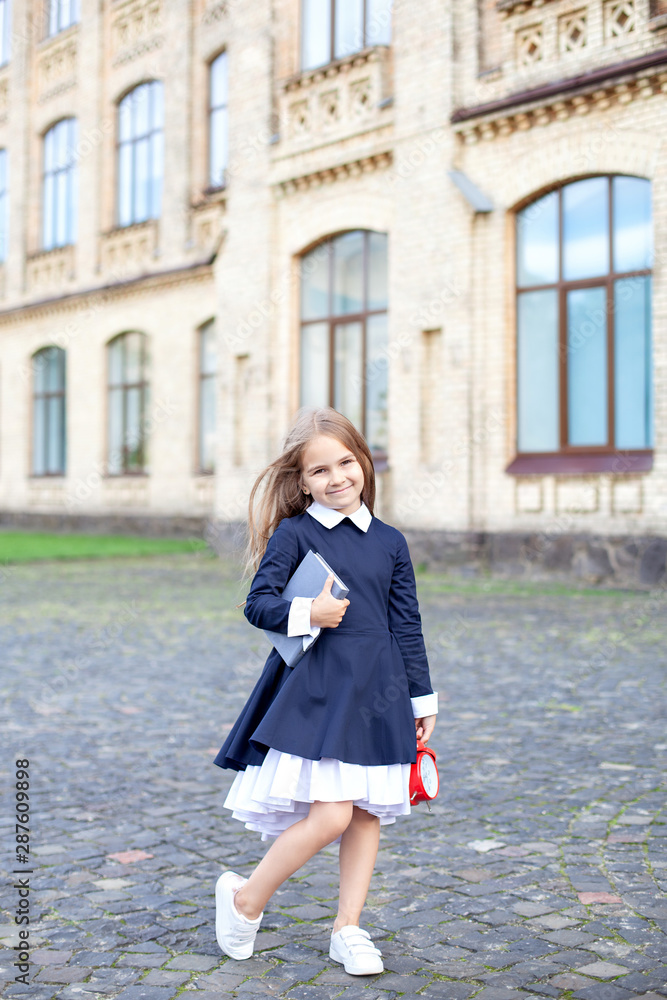 Back to school. Smart schoolgirl holds books and an alarm clock, ready for the new school year against the backdrop of the school. place to copy. Education concept. Primary school student. autumn