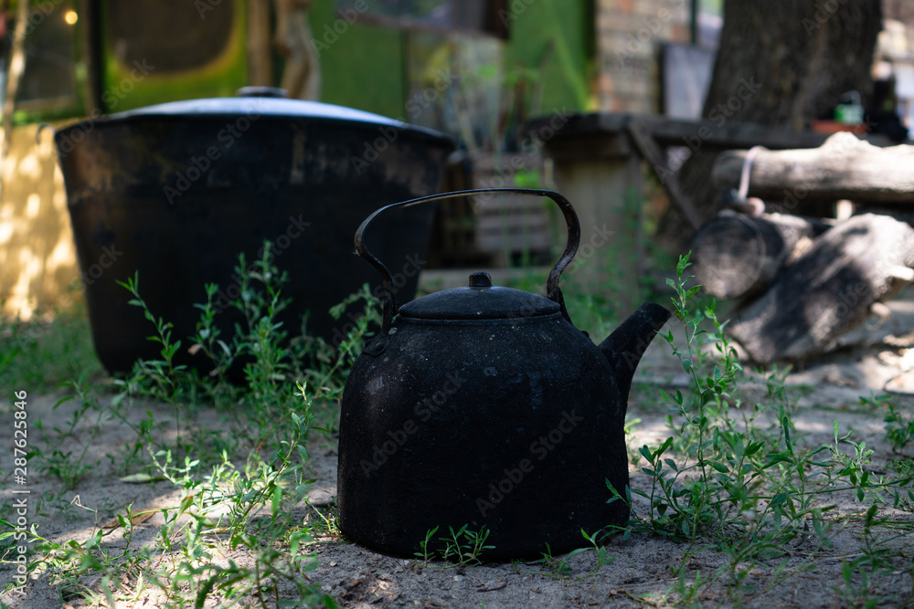 Old teapot for tourism. Black fired. Standing on green grass. on the background is a black burnt tourist pan. Dark tone. Horizontal orientation Old teapot for tourism. Black fired. Standing on green grass. on the background is a black burnt tourist pan. Dark tone. Horizontal orientation
