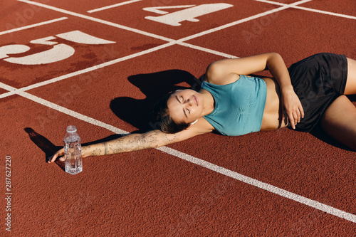 Beautiful athlete girl in sportswear dreamily lying on runner track with bottle after workout on city stadium photo