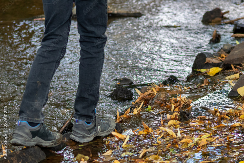 Junge mit schwarzer Hose und Wanderschuhen überquert einen Fluss über einen kleinen Damm aus Steinen und balanciert über glitschige Steine und Blätter und bekommt nasse Füße aber hat draußen viel Spaß Junge mit schwarzer Hose und Wanderschuhen überquert einen Fluss über einen kleinen Damm aus Steinen und balanciert über glitschige Steine und Blätter und bekommt nasse Füße aber hat draußen viel Spaß