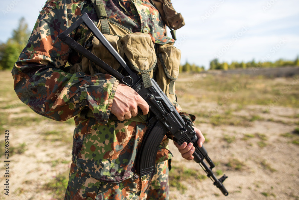 Fototapeta premium Soldier holds a machine gun in hands, close-up. Man in green camouflage on the battlefield with a black assault rifle