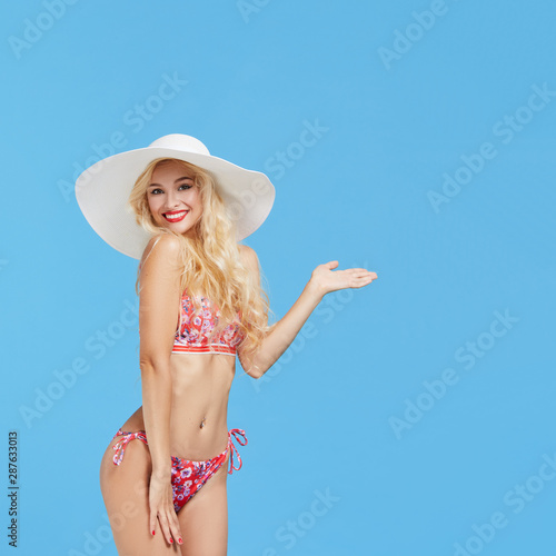 Portrait of a smiling and shocked attractive woman in bikini and hat points a hand to the right and up and looking at camera isolated over blue background. Travel and vacation concept. 