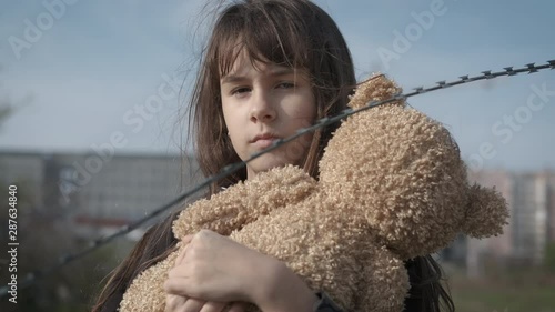 Homeless child with a teddy bear. Sad orphan girl with a bear behind a barbed wire. photo