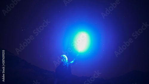 Silhouette of a man with blue light lamp in his hand standing against night starry sky. Long exposure photography.