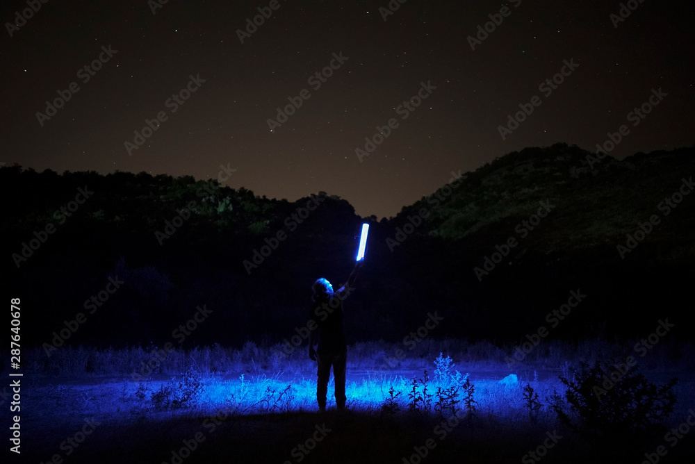 Silhouette of a man with blue light lamp in her hand standing against night starry sky. Long exposure photography.