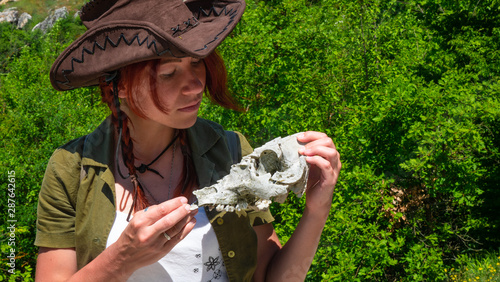 Εκτύπωση καμβά A beautiful archaeologist girl in a cowboy hat examines the skull of a domestic pig or wild boar near the settlements of the ancient people of cave cities