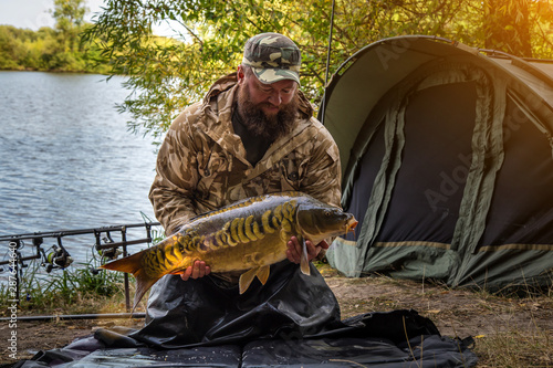 Fishing adventures, carp fishing. Mirror carp (Cyprinus carpio), freshwater fish. Angler with a big carp fishing trophy.Sunrise with a Carp Angler overlooking Lake photo