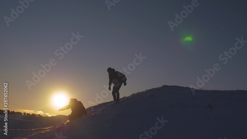 Silhouettes of men descending from high snowy mountain seeking adventure in glare of setting sun in winter. Concept of climbers who conquered mountain.Teamwork. Friends visiting terrain of mountains.