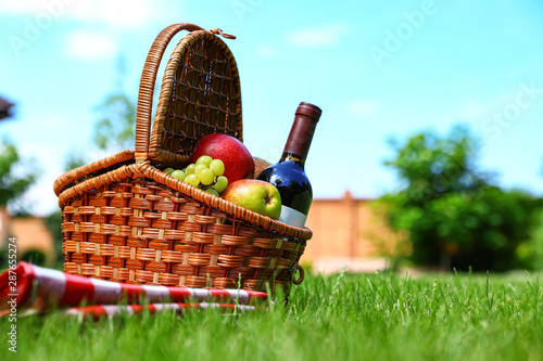 Picnic basket with fruits, bottle of wine and checkered blanket on green grass in garden. Space for text