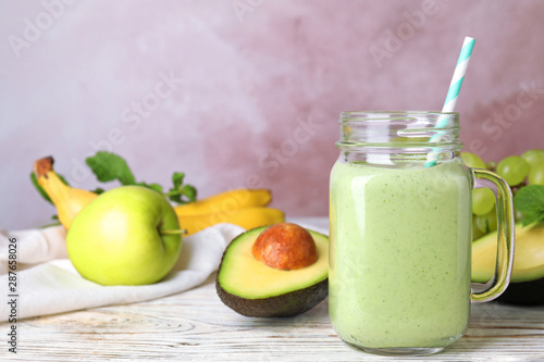 Mason jar of tasty smoothie and avocado on white table against pink background