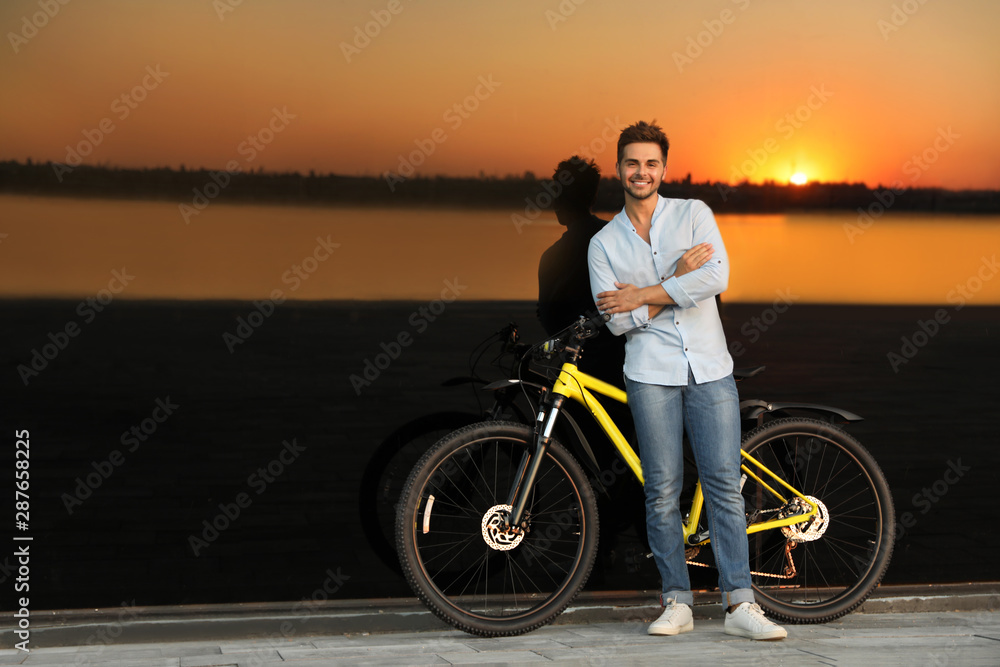Fototapeta premium Handsome young man with bicycle near glass building at sunset. Space for text