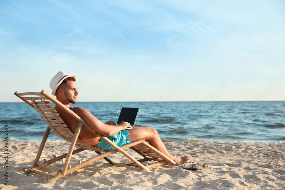 Young man with laptop in deck chair on beach