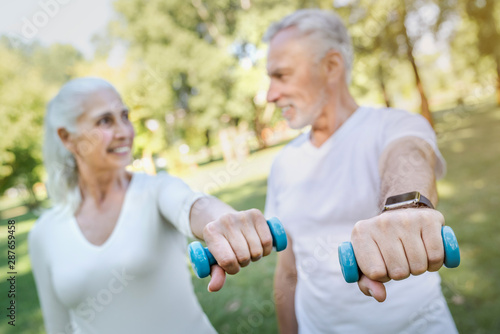 Senior couple is doing sport and fitness outdoors in park while looking on each other, focus on dumbbells