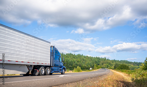 Big rig blue bonnet semi truck going with refrigerator semi trailer on the road with forest and meadows