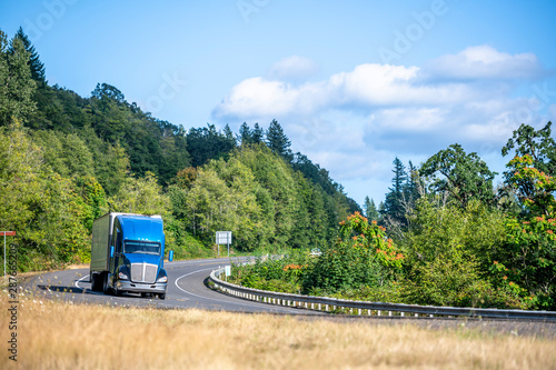 Big rig bonner blue semi truck with semi trailer transporting cargo running on the winding road with green forest trees photo