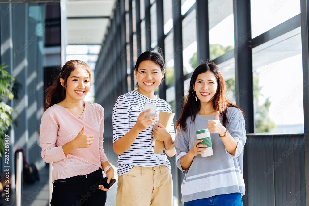 Smart smiling Asian women with notebooks and coffee gesturing thumb up and looking at camera in corridor Smart smiling Asian women with notebooks and coffee gesturing thumb up and looking at camera in corridor