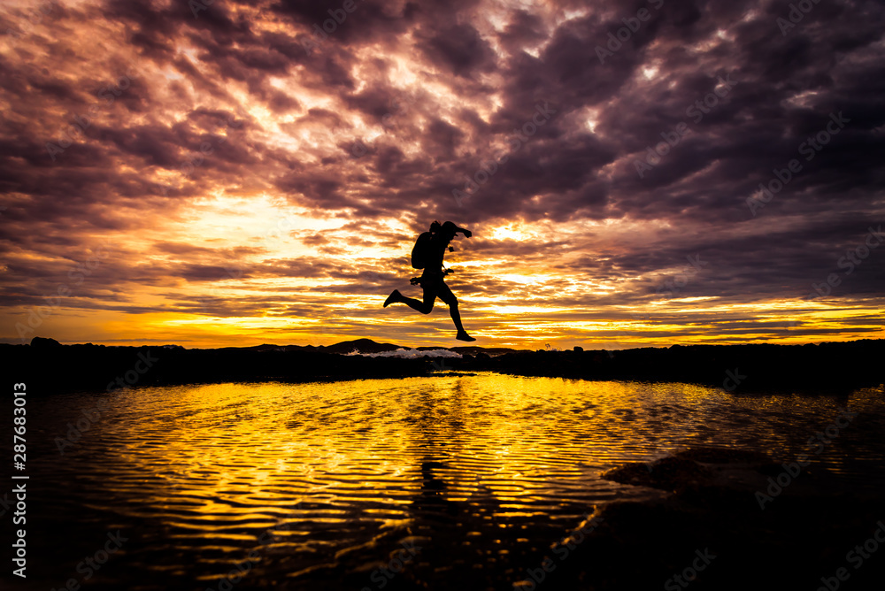Silhouetted man running and jumping  across water at sea with dark sky ,black cloud at sunset time background.