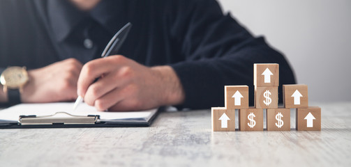 Man writing in document. Wooden cubes on the desk with arrows and dollar signs